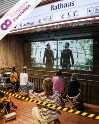 Menschen sitzen bei einer Theateraufführung von Lokstoff an einer Bahnstation am Rathaus in Stuttgart mit Silent Kopfhörern von Headphone Revolution.