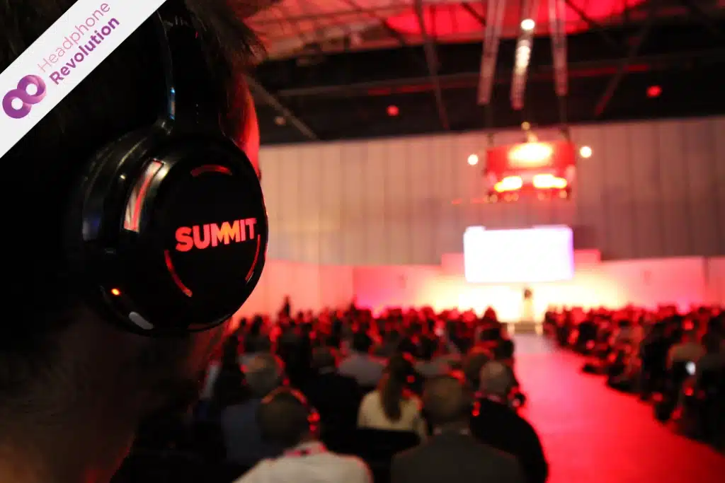 Ein Mann, der einen Silent Conference Kopfhörer SCF mit Logo Branding von Adobe Summit trägt und auf einer Messe auf eine Bühne schaut und gleichzeitig dem mit vielen anderen Menschen einer Rednerin zuhört. Menschen bei der Adobe SUMMIT Conference tragen Silent Kopfhörer mit gebrandeten Seitenteilen.