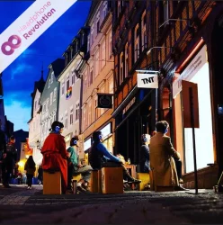 Menschen sitzen in einer Altstadt vor einem Schaufenstertheater und schauen einer Theatervorstellung zu, bei der der Ton über ein spezielles Silent Kopfhörer System übertragen wird.