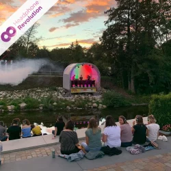 Menschen sitzen bei einem Silent Disco Festival am Ufer und empfangen den Ton einer Band, die gerade ein Konzert spielt, über Neon Lights Superior Kopfhörer.