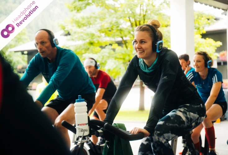 Sportliche Menschen auf Spinningrädern trainieren Outdoor und hören dabei die gleiche Musik über Neon Lights BS Plus+ Kopfhörer von Headphone Revolution.