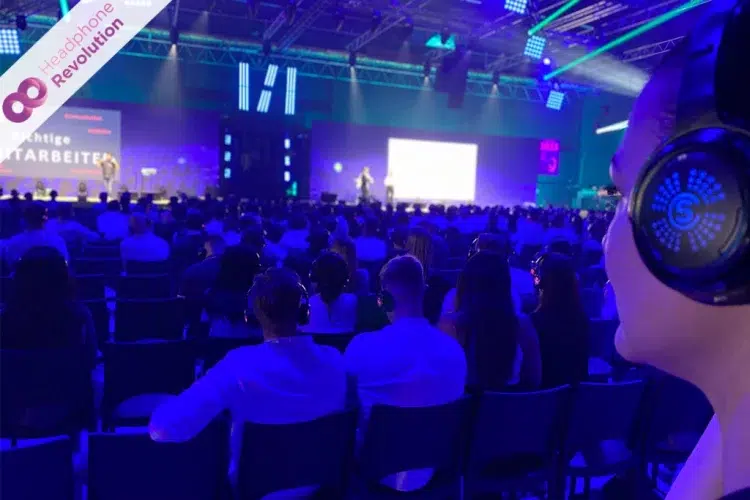 Ein großes Publikum sitzt beim MHP Festival vor zwei Bühnen während zwei verschiedene Speaker gleichzeitig einen Vortrag halten und dabei das Silent Conference Kopfhörersystem "SCF", auf dem der Ton übertragen wird, tragen.