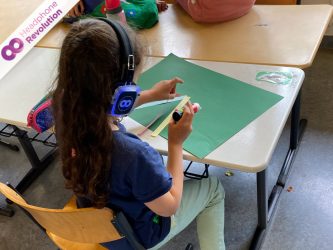 Ein Kind das an einem Tisch bastelt und dabei Kopfhörer trägt, auf dem die Bastelanleitung übertragen wird.