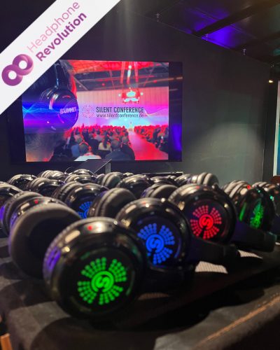 Einige verschieden farbig leuchtende Silent Conference Kopfhörer liegen aufgereiht auf einem Tisch auf einer Messe, bei dem im Hintergrund ein Video läuft.