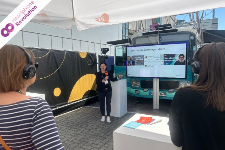 Eine ZDF Moderatorin hält eine Präsentation an ihrem Stand, bei dem die Teilnehmer und Teilnehmerinnen Silent Conference Kopfhörer tragen, um den Ton besser zu verstehen.