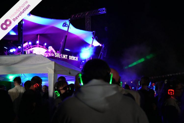 Menschen feiern vor der Bühne auf einem Festival, auf welchem DJs auflegen und tragen dabei Silent Disco Kopfhörer auf die die Musik übertragen wird.