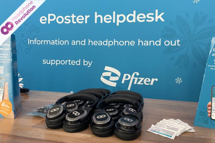 Ein paar HRT12PRO Kopfhörer liegen auf einen Tisch, hinter dem ein Pfizer Plakat hängt