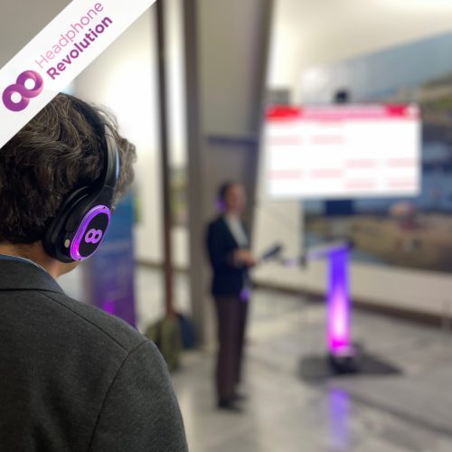 Während der Sprecher einer Silent Conference im Hintergrund einen Vortrag hält, trägt ein Besucher HRT12PRO Kopfhörer